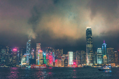 Illuminated modern buildings by bay against sky at night