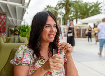 Pretty girl with long hair sitting in bar outdoor in city, drinking lemonade on straw