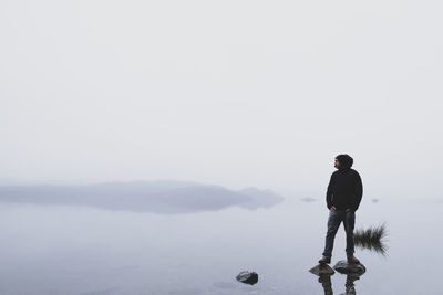 Man standing on rocks in lake against clear sky during foggy weather