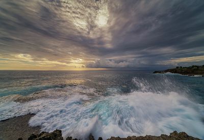 Waves rushing on shore against cloudy sky during sunset