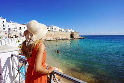 Back view of young woman with hat and dress enjoying seascape in monopoli town, bari, italy.