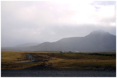 Distant view of road amidst field by mountain against cloudy sky