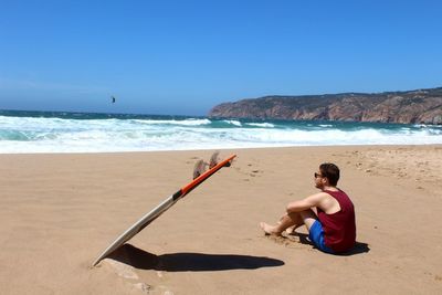 Side view of young man with surfboard sitting at beach against blue sky during sunny day