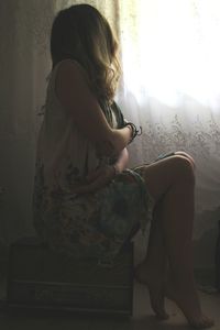 Side view of woman sitting on box by window at home