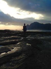 Rear view of man standing on beach against cloudy sky