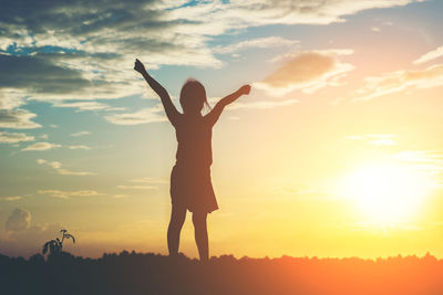 Rear view of silhouette girl standing against sky on field during sunset