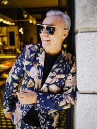 Happy male wearing stylish suit with floral print leaning on stone wall of building and looking away