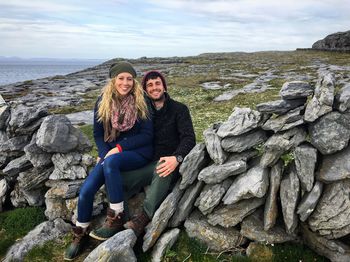 Portrait of smiling man and woman sitting on rocks against cloudy sky