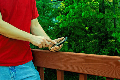 Midsection of man using mobile phone while standing by railing and trees