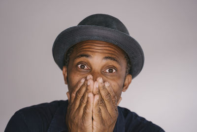 Man wearing hat covering mouth with hands against gray background