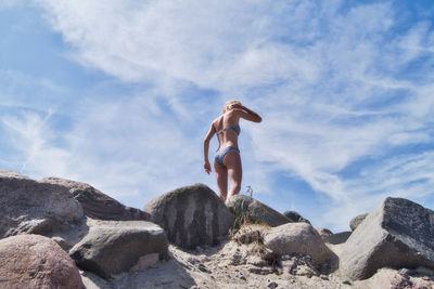Low angle view of young woman in bikini standing on rocks against sky