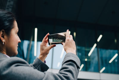 Side view of businesswoman photographing modern building with smart phone while standing in city