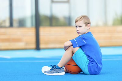 Portrait of boy sitting with basketball on sports court