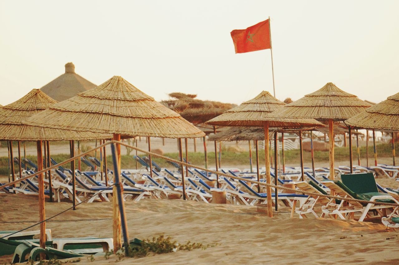 clear sky, beach, sand, beach umbrella, built structure, sky, parasol, sea, flag, copy space, day, tranquil scene, tranquility, chair, sunshade, absence, in a row, thatched roof, wood - material, water