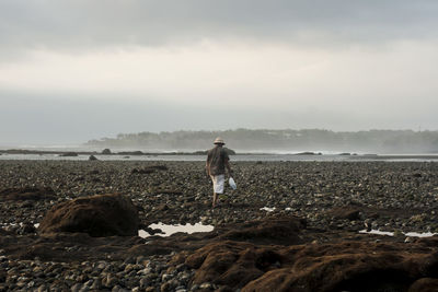 Rear view of man walking at beach against cloudy sky