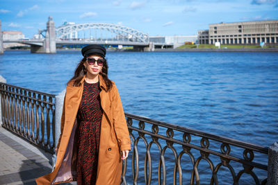 Young woman in sunglasses posing on embankment in brown coat on spring sunny day