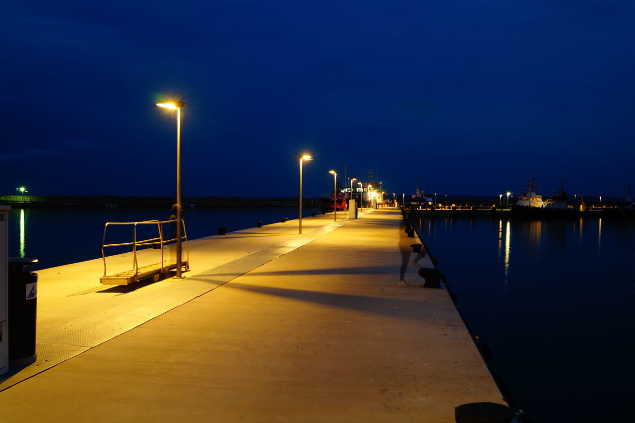 street light, illuminated, water, blue, reflection, architecture, lake, building exterior, sea, in a row, pier, calm, the way forward, ocean, walkway, long, cloud, lamp post, bridge, harbor, promenade, sky, tranquil scene