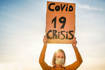 Portrait of woman wearing mask holding banner against sky at sunset