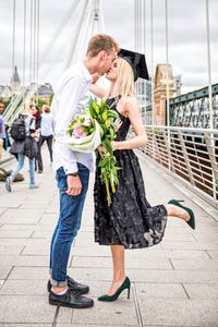 Woman wearing mortarboard and holding bouquet kissing boyfriend on bridge