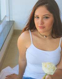 Portrait of woman holding rose while sitting on sheet at home