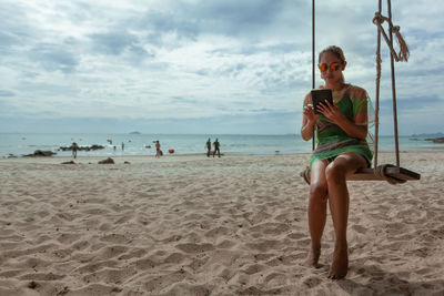 Full length of woman using digital tablet while sitting on swing at beach against sky