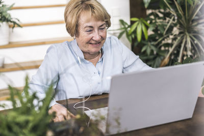 Happy blond senior woman watching movie on laptop at table in living room