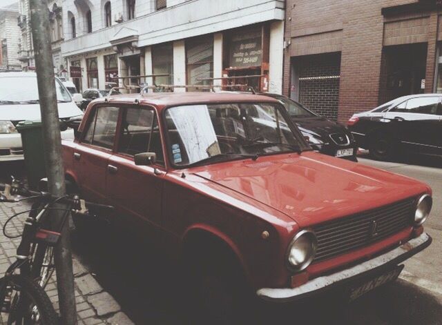 land vehicle, car, transportation, mode of transport, building exterior, architecture, built structure, street, vintage car, city, stationary, travel, road, old-fashioned, parking, damaged, red, day, vehicle hood, city life, outdoors, in front of, obsolete, parked, no people, vibrant color, motor vehicle