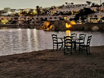 Chairs and table by river against buildings in city at dusk