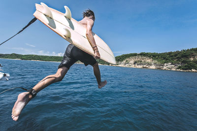 Man with surfboard jumping into sea against sky during sunny day