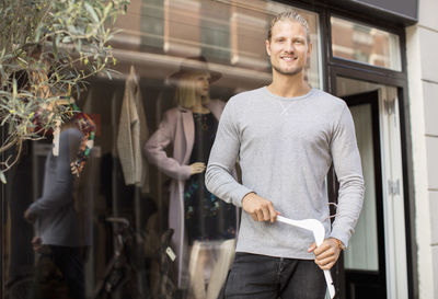 Portrait of smiling owner holding coathanger while standing outside clothing store