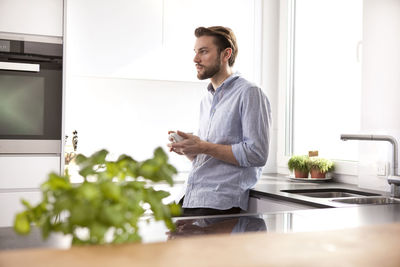 Pensive young man with cup of coffee standing in his kitchen