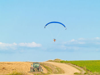 Person powered paragliding over agricultural field against blue sky