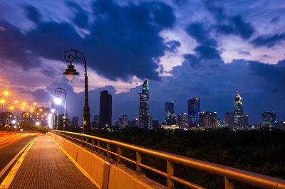 Illuminated street in city against cloudy sky at night