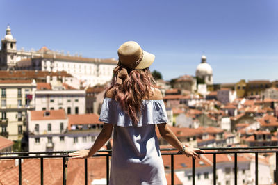 Rear view of young woman looking at buildings while standing by railing in balcony against clear sky