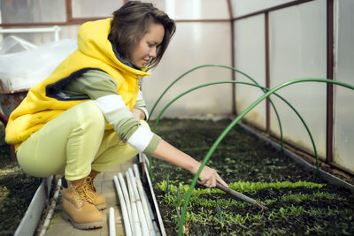 Side view of woman loosening soil with gardening fork while crouching in greenhouse