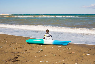 Rear view of man sitting on shore at beach against sky