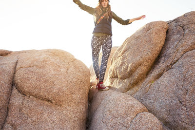 Low angle view of playful woman with arms outstretched walking on rock against sky