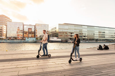 Happy couple riding electric push scooters on promenade by river in city against sky