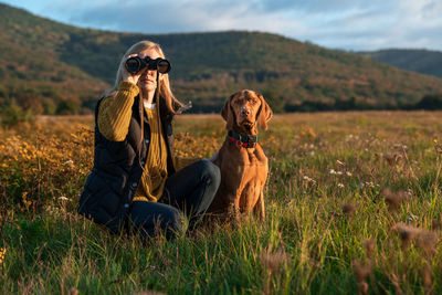 Young female hunter using binoculars for bird spotting with hungarian vizsla dog by her side.