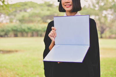 Midsection of young woman in graduation gown holding file while standing at park