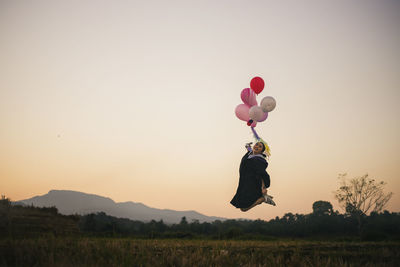 Full length of woman flying balloons against sky during sunset