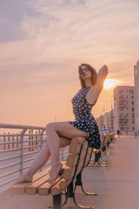 Young woman sitting on sunglasses against sky during sunset
