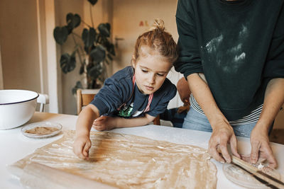 Girl sprinkling cinnamon powder on dough while helping mother in kitchen