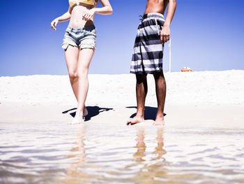 Low section of friends standing on shore at beach against clear sky