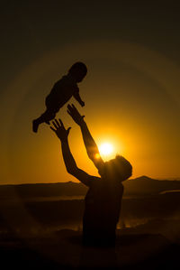 Silhouette father throwing son in air against sky during sunset