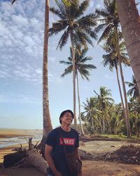 Portrait of young man standing by palm trees on beach against sky