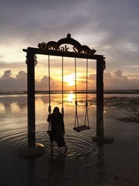 Woman sitting on swing over sea against cloudy sky during sunset