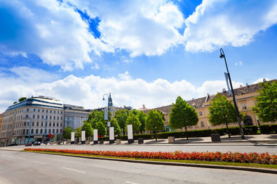 Road by buildings against cloudy sky