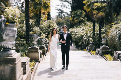 Happy lovers the bride and groom in wedding outfits walk among plants and palm trees in the old park