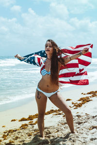 Woman holding american flag at beach against sky